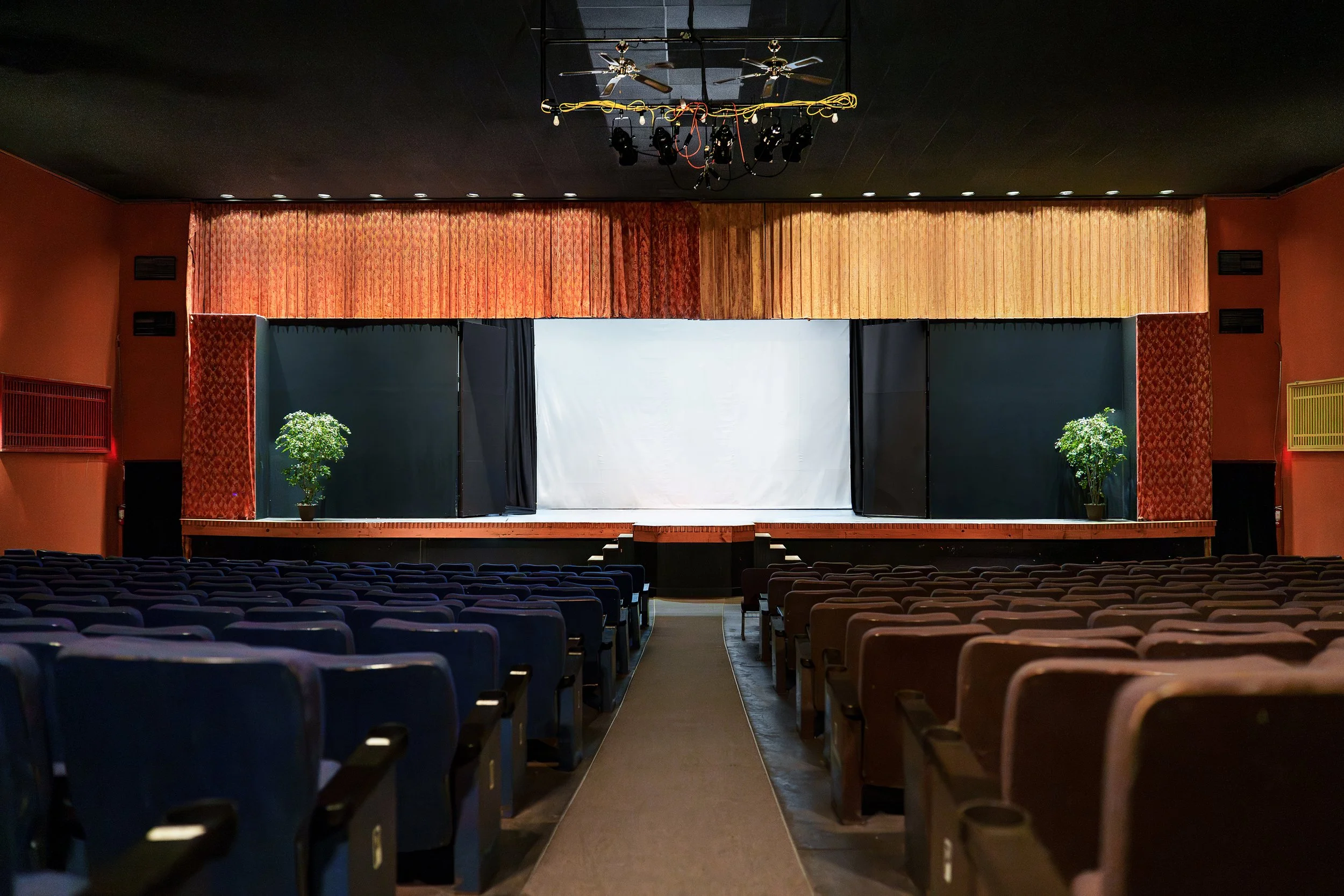 Empty auditorium with a stage, black curtains, potted plants on each side, and overhead lighting and equipment.