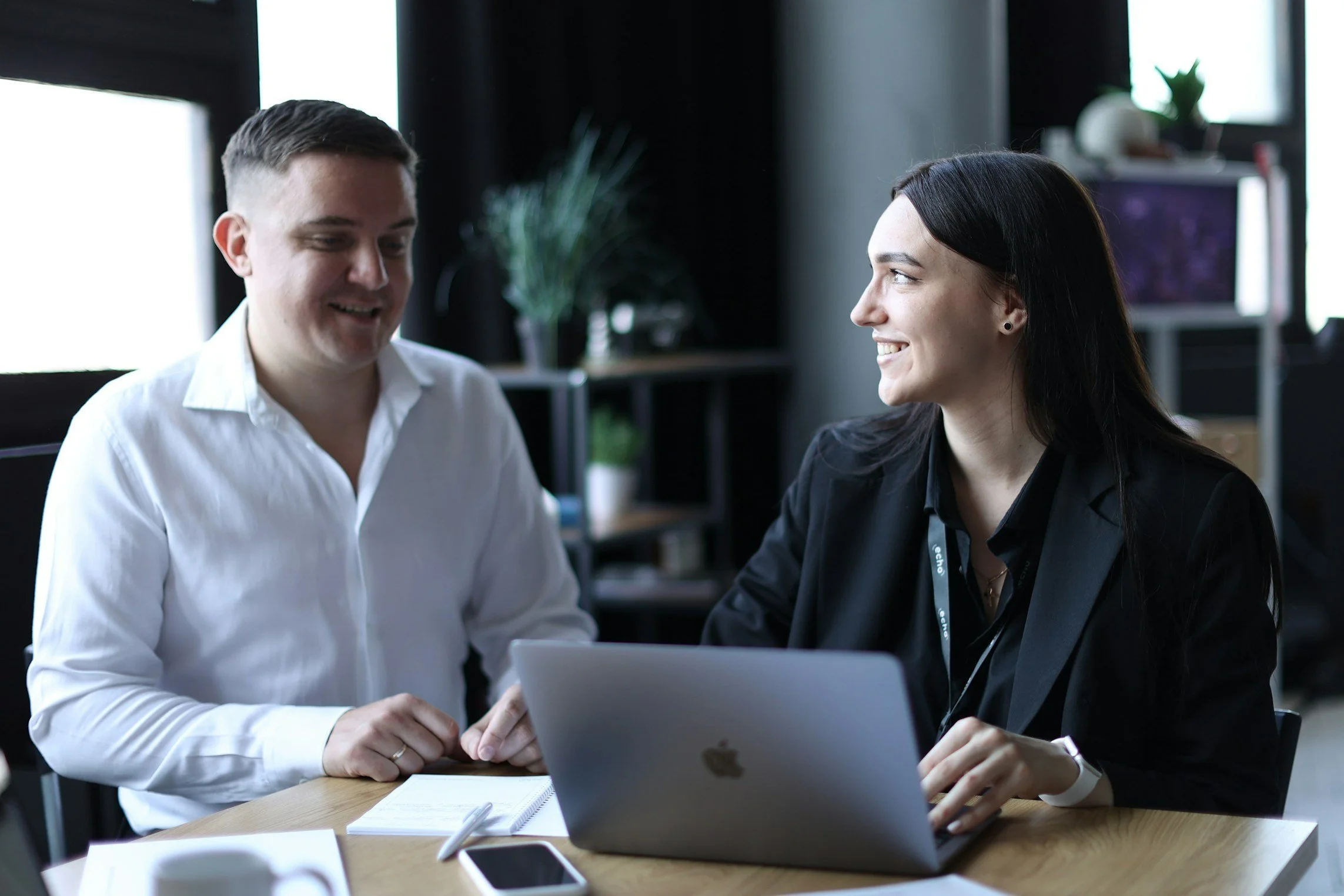 Two people having a meeting at a desk with a laptop and notepad in an office setting.