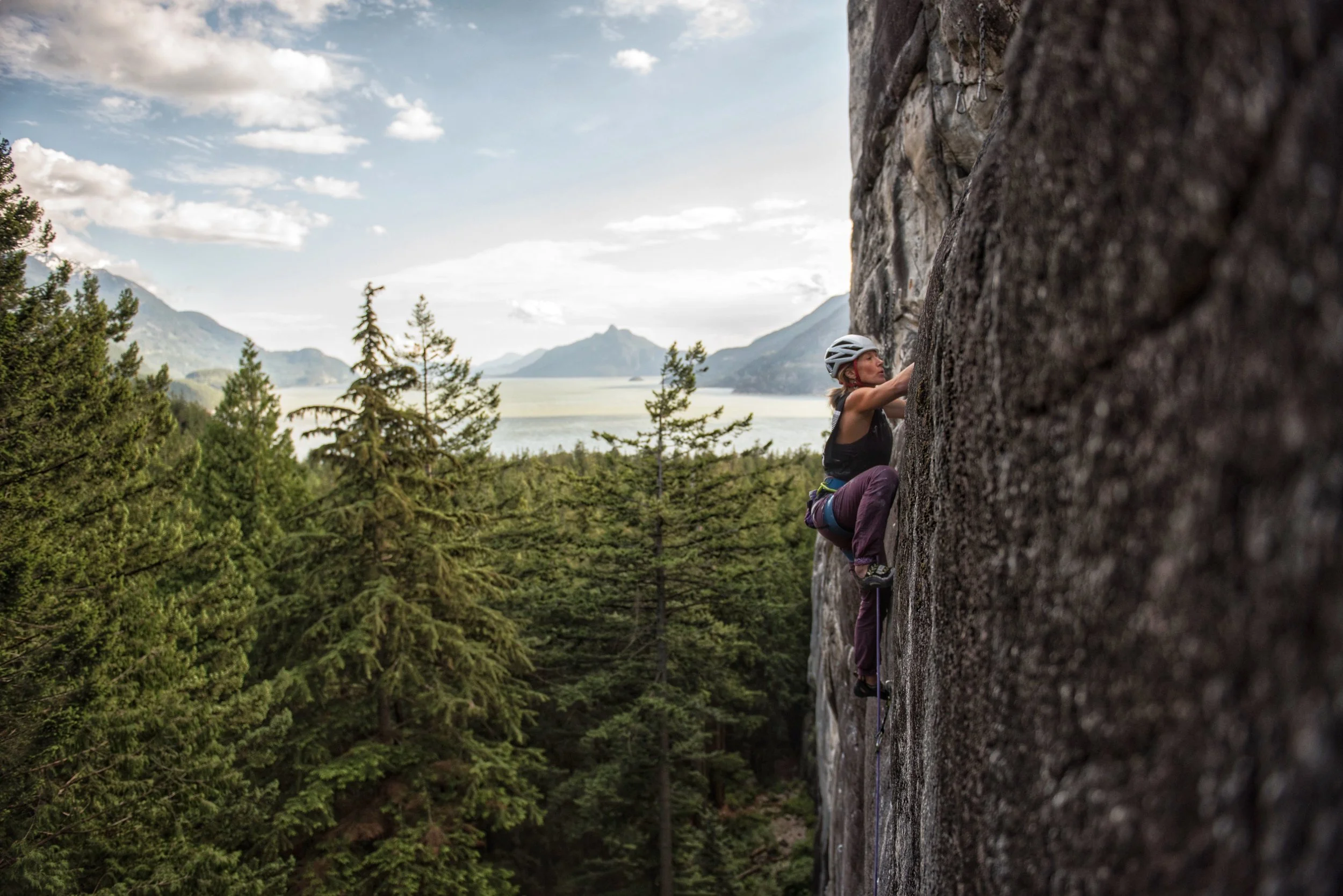 A woman climbing a rock face in a mountainous forest with a lake and mountains in the background.
