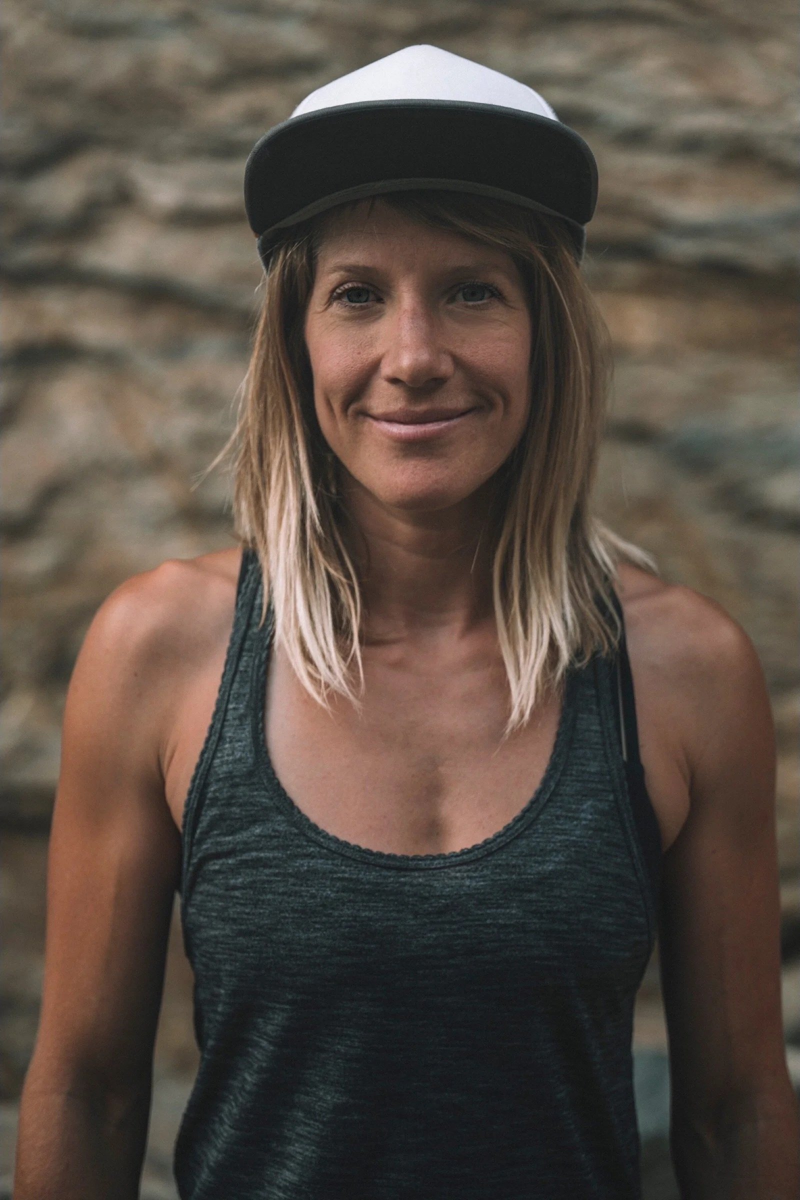 A woman in a black tank top and a black and white cap, smiling at the camera, standing against a stone wall background.