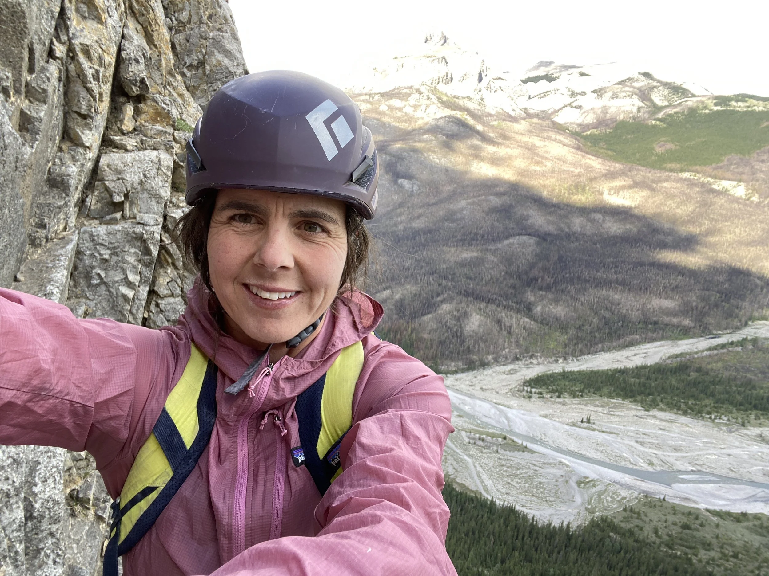 A woman wearing a helmet and pink jacket taking a selfie on a rocky mountain trail with a forested valley and snow-capped mountains in the background.