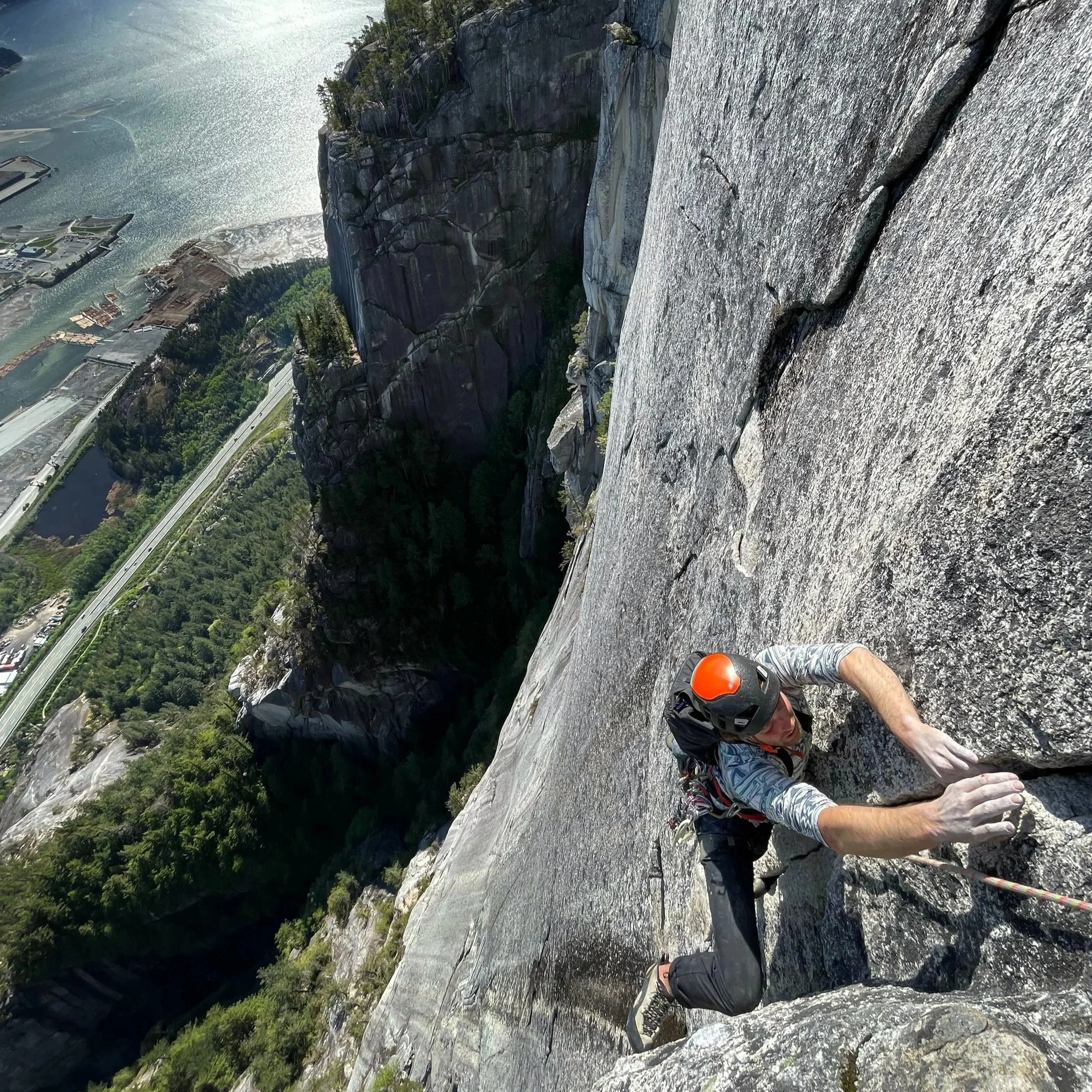 A person rock climbing up a steep granite cliff with a view of a valley, trees, water, and buildings below.