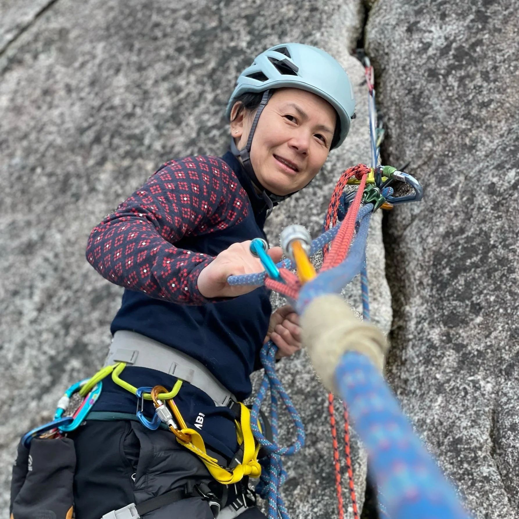 A woman rock climbing outdoors on a gray granite rock face, wearing a gray helmet, a black and red patterned long-sleeve shirt, and climbing gear including carabiners and ropes.