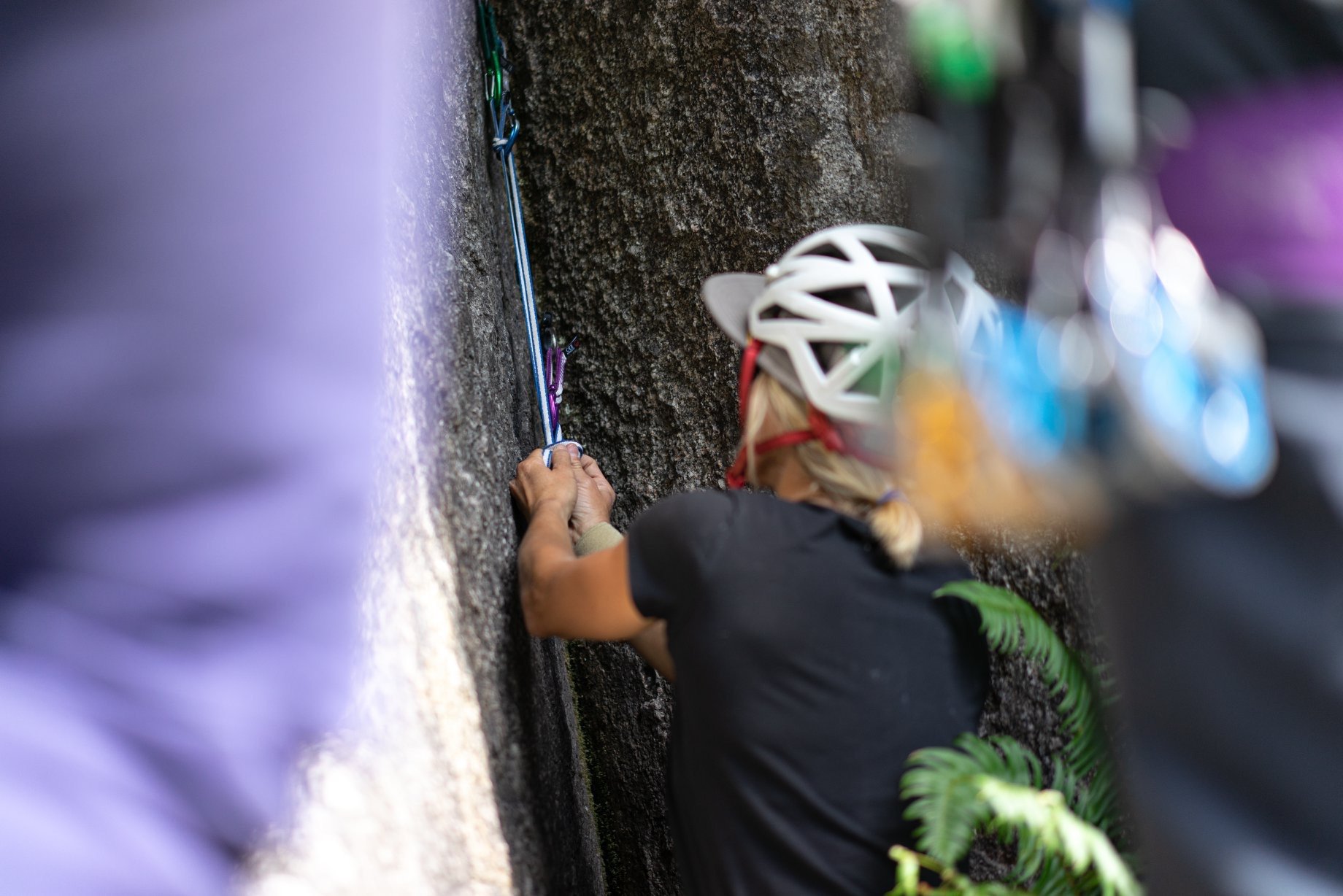 A woman rock climbing outdoors, wearing a white helmet and gripping climbing holds on a vertical rock wall.