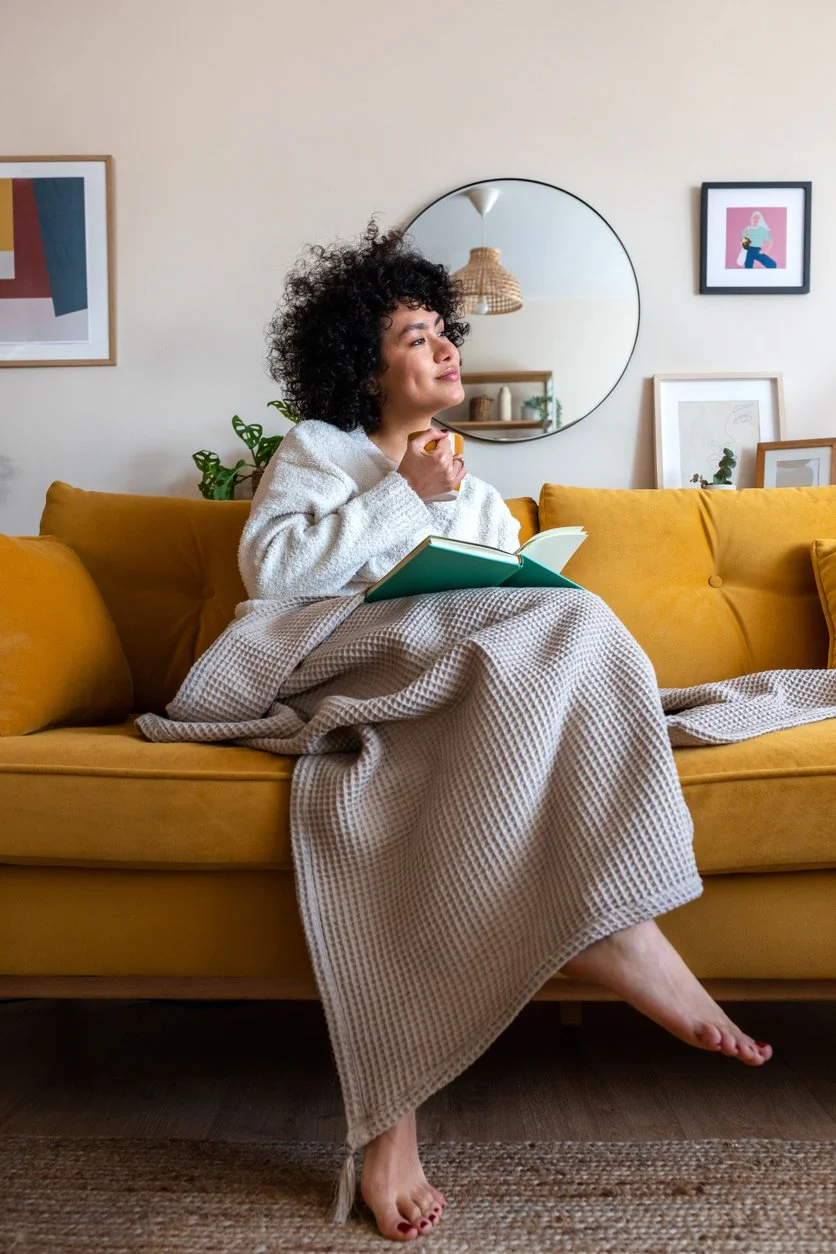 A woman rests on a mustard-yellow couch with a book and a blanket.