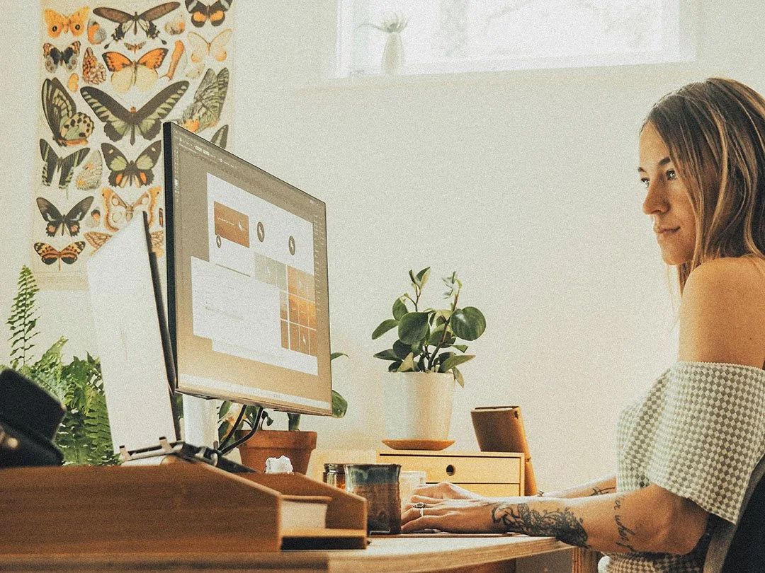 A woman working on a computer at a desk with plants and butterfly posters on the wall.