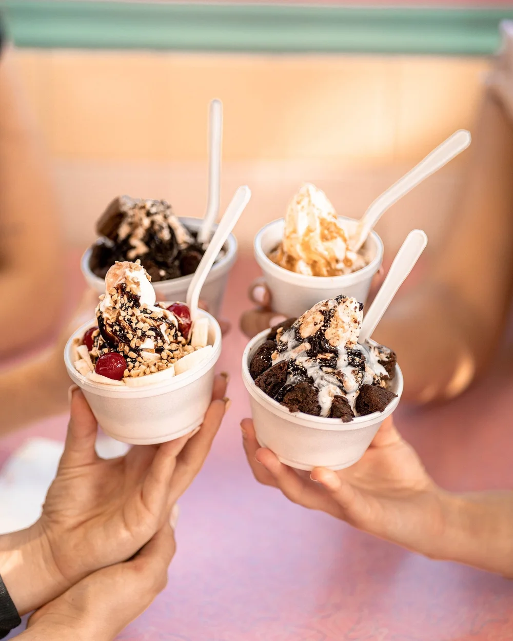 Four sundaes in white cups with spoons, topped with brownie pieces, chocolate and white drizzles, caramel, banana, cherries and chopped nuts, raised together in a toast, at Merla-Mae Ice Cream in London, Ontario