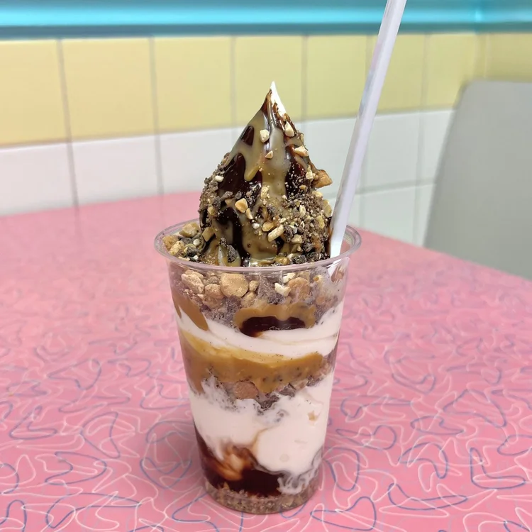 Peanut butter parfait in a clear cup with layers of soft-serve, peanut butter sauce, fudge and crushed peanuts, on a pink retro table in front of pastel tiles at Merla-Mae Ice Cream in London, Ontario