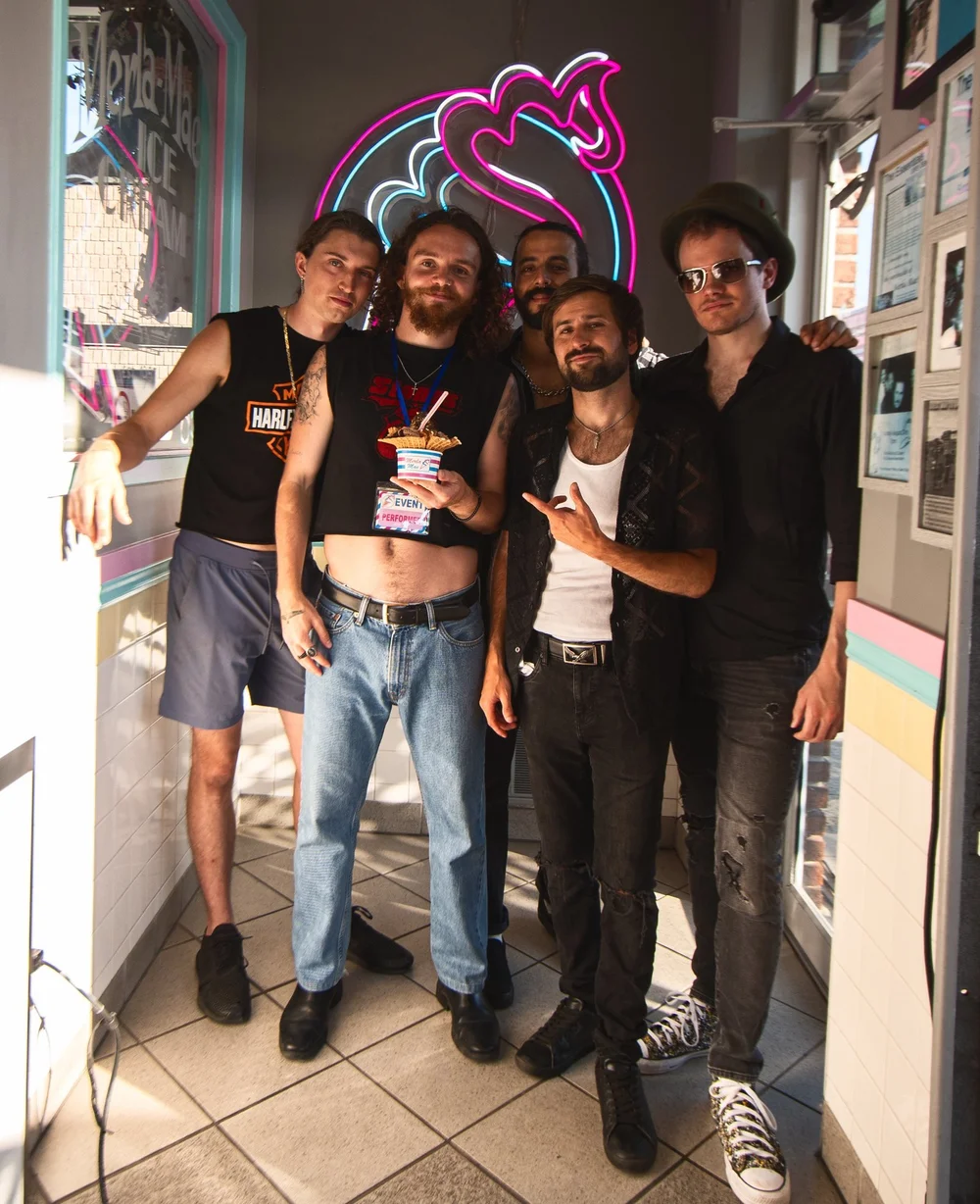 Tyrone Traher and The Rottweilers posing together under the neon cone sign inside Merla-Mae Ice Cream in London, Ontario, at National Ice Cream Day 2025