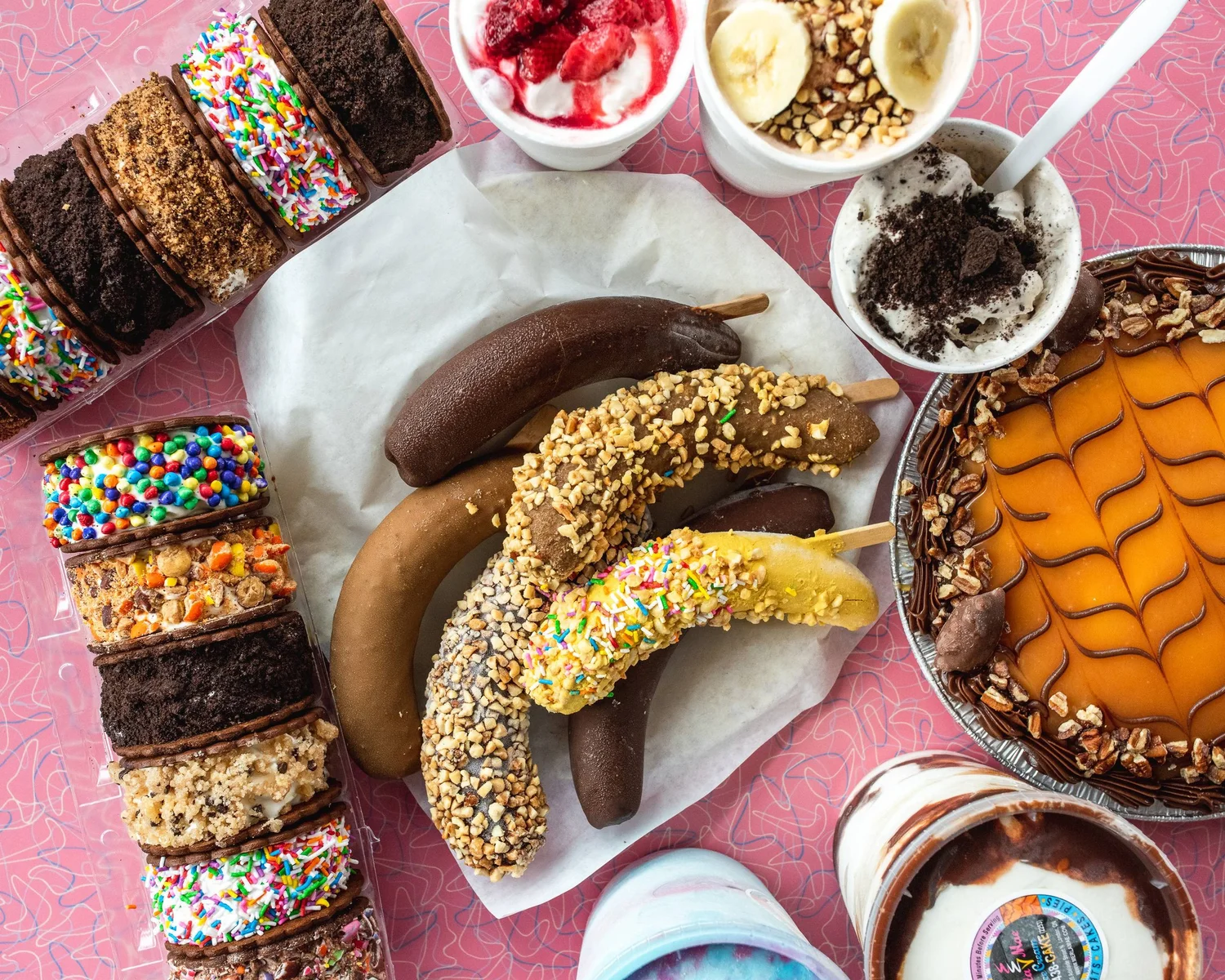 Take-home frozen treats on a pink retro tablecloth: ice cream sandwiches rolled in sprinkles, candy and cookie crumbs, chocolate-dipped banana dip sticks with nuts and sprinkles, topped cups, a pie and take-home tubs at Merla-Mae Ice Cream in London, Ontario