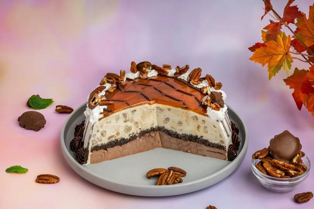 Turtle ice cream cake cut open to show its pecan, cookie crumb and chocolate layers, topped with caramel and pecans beside autumn maple leaves, from Merla-Mae Ice Cream in London, Ontario