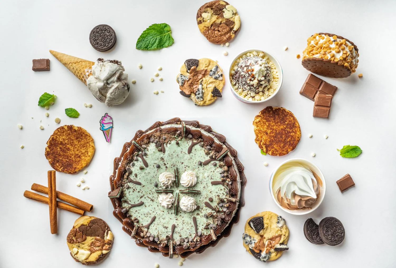 Flat lay of a mint-green ice cream cake with chocolate drizzle and whipped cream rosettes, surrounded by a waffle cone, cookies, cups of soft-serve, cinnamon sticks, chocolate squares and mint leaves on a white table, at Merla-Mae Ice Cream in London, Ontario