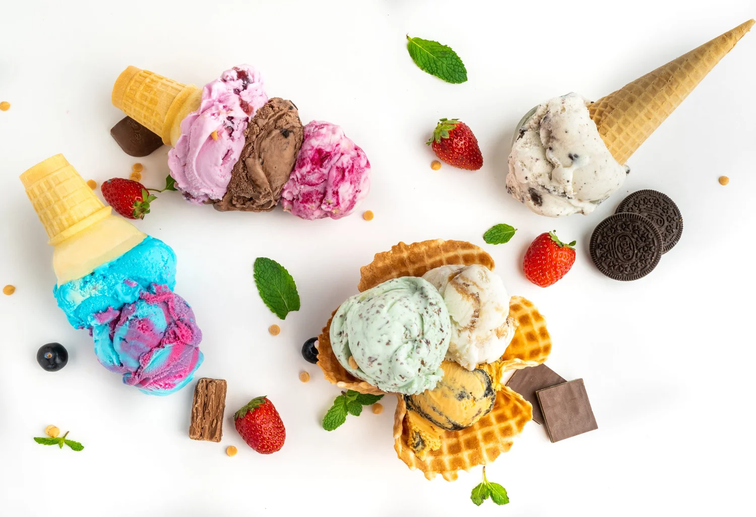 Hard scoop ice cream on a white table: pink, chocolate and berry scoops on a cake cone, blue and purple swirl scoops on another cone, a cookie-studded scoop on a sugar cone and three scoops in a waffle dish, with strawberries, cookies and mint, at Merla-Mae Ice Cream in London, Ontario