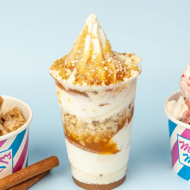 Soft-serve parfait layered with caramel sauce and crumble in a clear cup between two Merla-Mae cups at Merla-Mae Ice Cream in London, Ontario