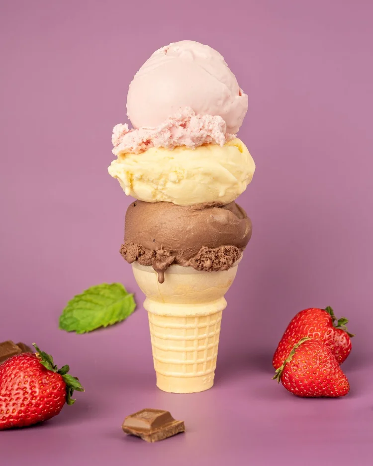 Three scoops of hard ice cream in pale pink, pale yellow and chocolate stacked on a cake cone at Merla-Mae Ice Cream in London, Ontario