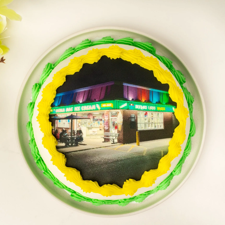 Round portrait ice cream cake with a photo of the lit-up ice cream shop at night on top, framed by yellow and green piped borders, at Merla-Mae Ice Cream in London, Ontario
