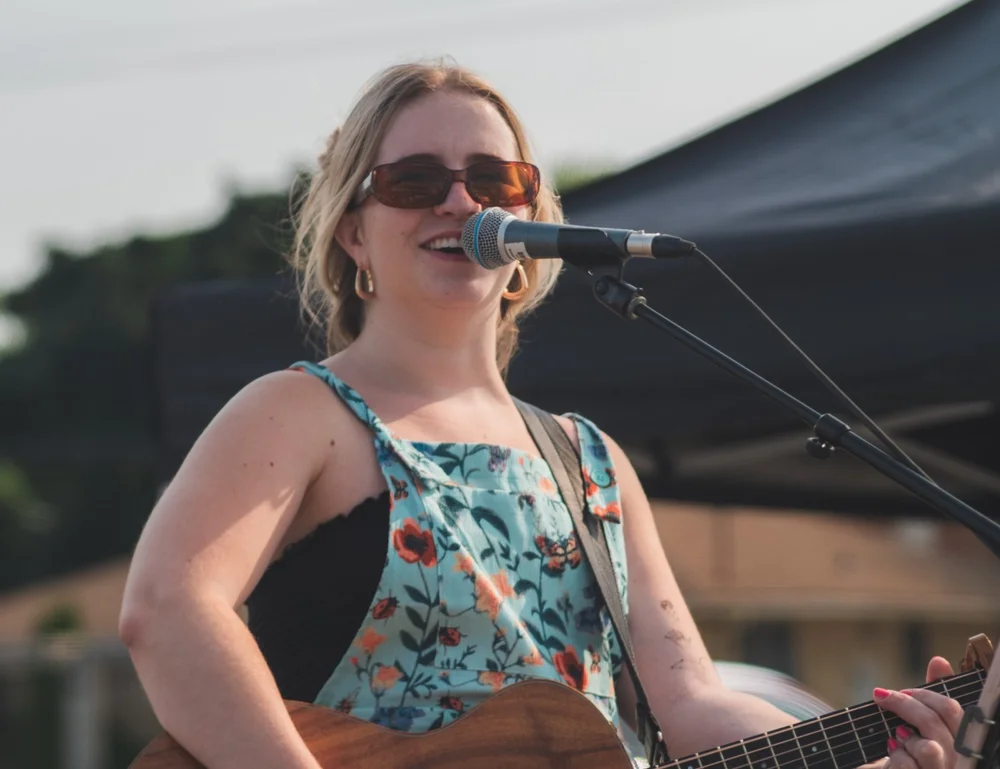 Sarina Haggarty smiling mid-song with her guitar at National Ice Cream Day 2024, Merla-Mae Ice Cream in London, Ontario