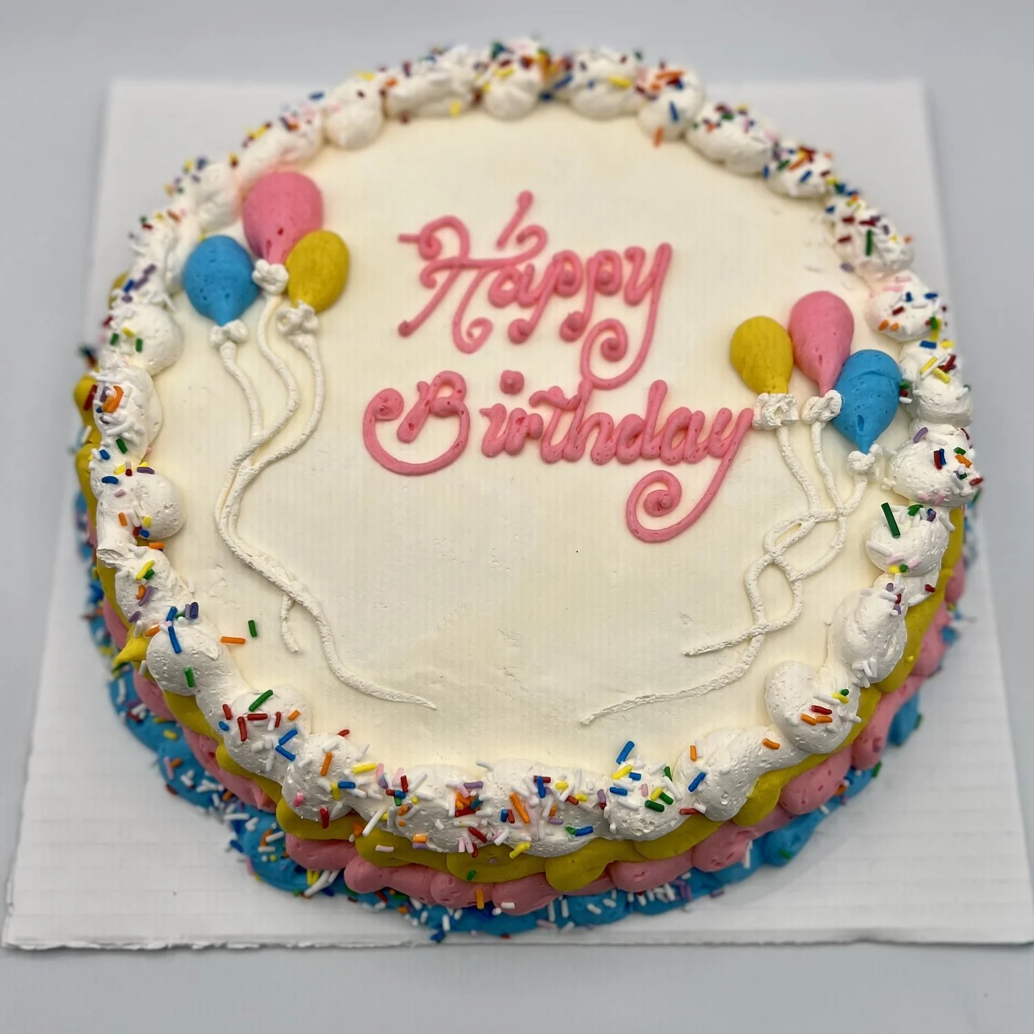Round ice cream cake with Happy Birthday piped in pink icing, blue, pink and yellow piped balloons, a sprinkle-topped whipped border and yellow, pink and blue piped sides, at Merla-Mae Ice Cream in London, Ontario