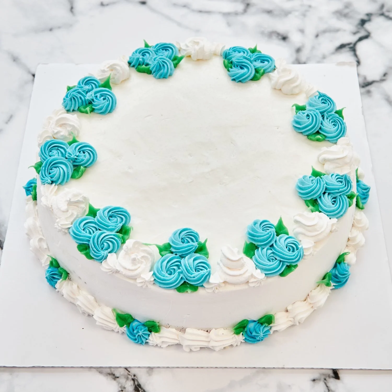 Round white ice cream cake with a smooth blank top, ringed with light blue and white piped rosettes and green leaves, with small blue rosettes on the bottom border, at Merla-Mae Ice Cream in London, Ontario