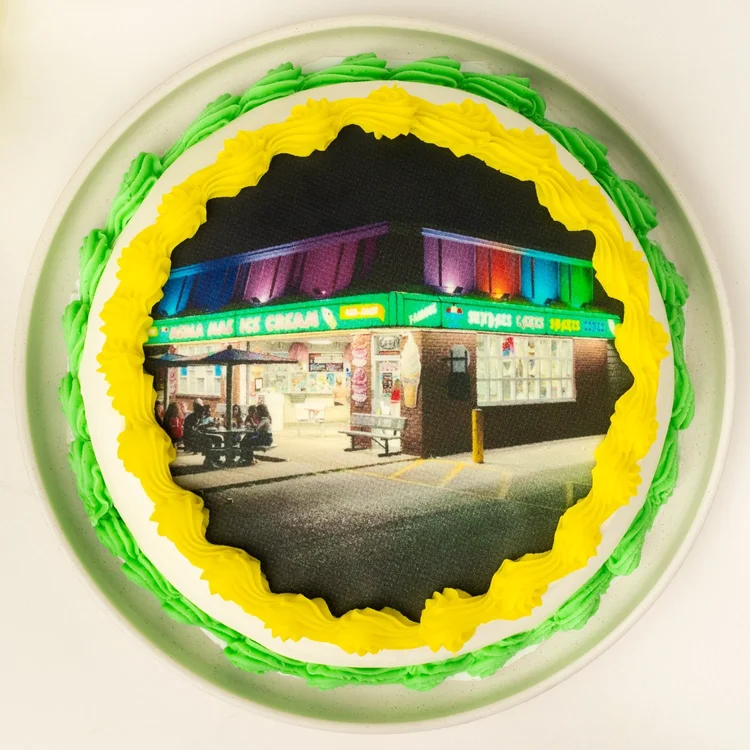 Round portrait ice cream cake seen from above, with an edible photo of the lit-up ice cream shop at night printed on top, framed by yellow and green piped borders, at Merla-Mae Ice Cream in London, Ontario