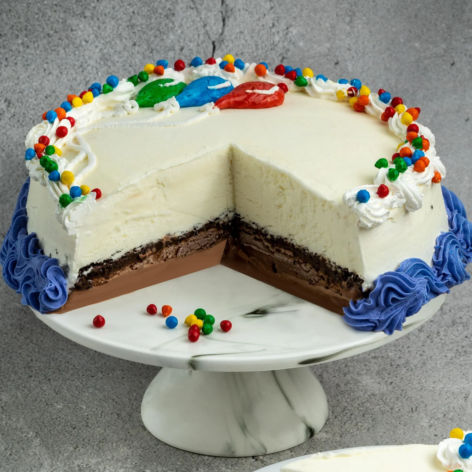 Ice cream cake cut open to show a chocolate ice cream base, a dark cookie crunch layer and vanilla ice cream on top, decorated with colourful candies, piped balloons and blue icing, on a marble cake stand at Merla-Mae Ice Cream in London, Ontario