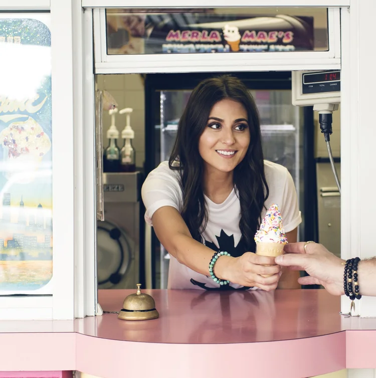A smiling server hands a rainbow-sprinkle soft-serve cone across the pink counter of the walk-up window at Merla-Mae Ice Cream in London, Ontario