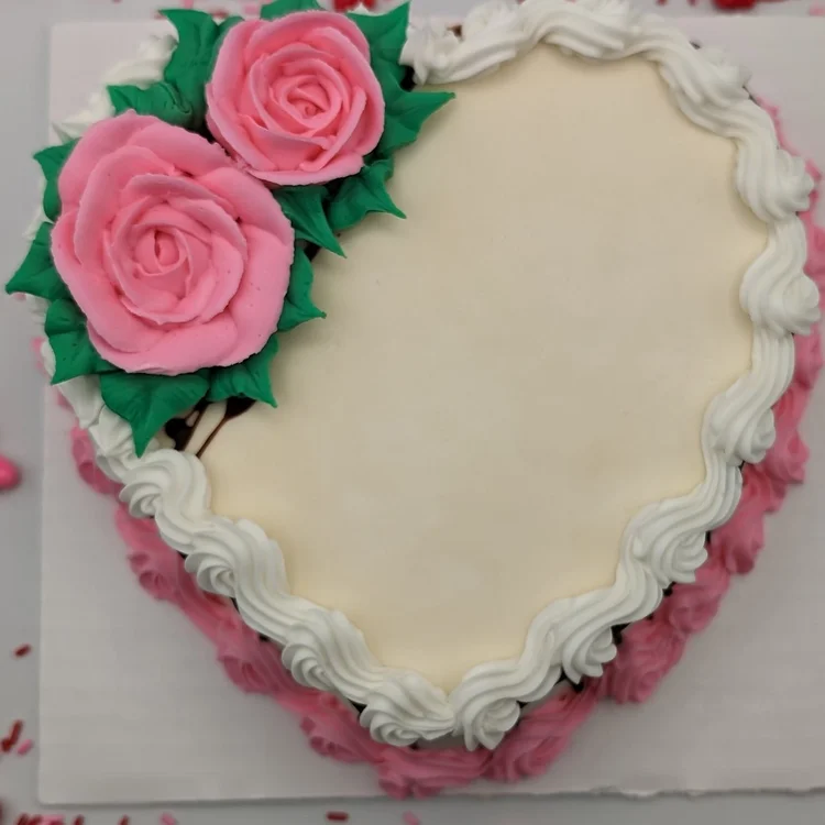 Heart-shaped ice cream cake seen from above, with a smooth ivory top, two large pink piped roses with green leaves, a white piped shell border and pink rosettes around the sides, at Merla-Mae Ice Cream in London, Ontario