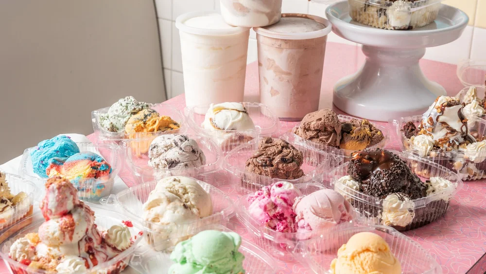 Scoops of ice cream and sundaes packed in clear takeout containers, with tall cups of ice cream behind, on a pink table at Merla-Mae Ice Cream in London, Ontario