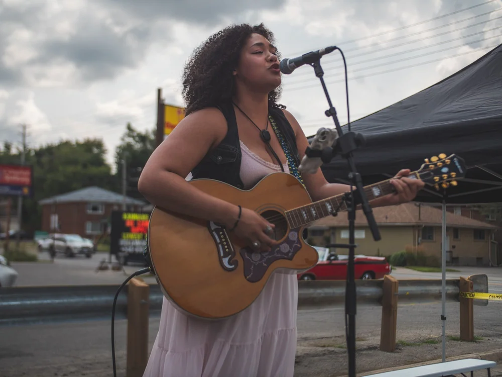 Gianna Rigopoulos singing with her acoustic guitar on the parking-lot stage at National Ice Cream Day 2024, Merla-Mae Ice Cream in London, Ontario