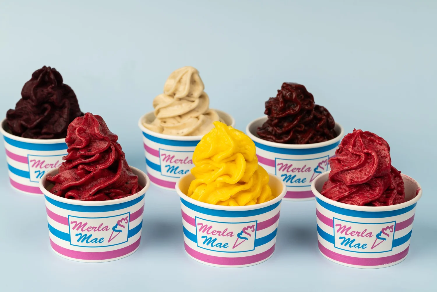 Six pink-and-blue striped cups of frozen yogurt and pressed fruit swirls in deep purple, red, cream and bright yellow on a pale blue backdrop at Merla-Mae Ice Cream in London, Ontario