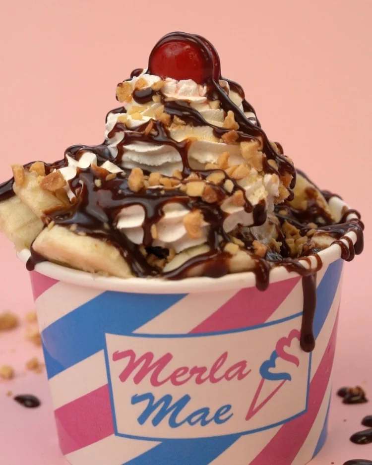 Sundae heaped with sliced bananas, whipped cream, chopped peanuts and a cherry, with chocolate fudge dripping over the rim of a striped Merla-Mae cup at Merla-Mae Ice Cream in London, Ontario
