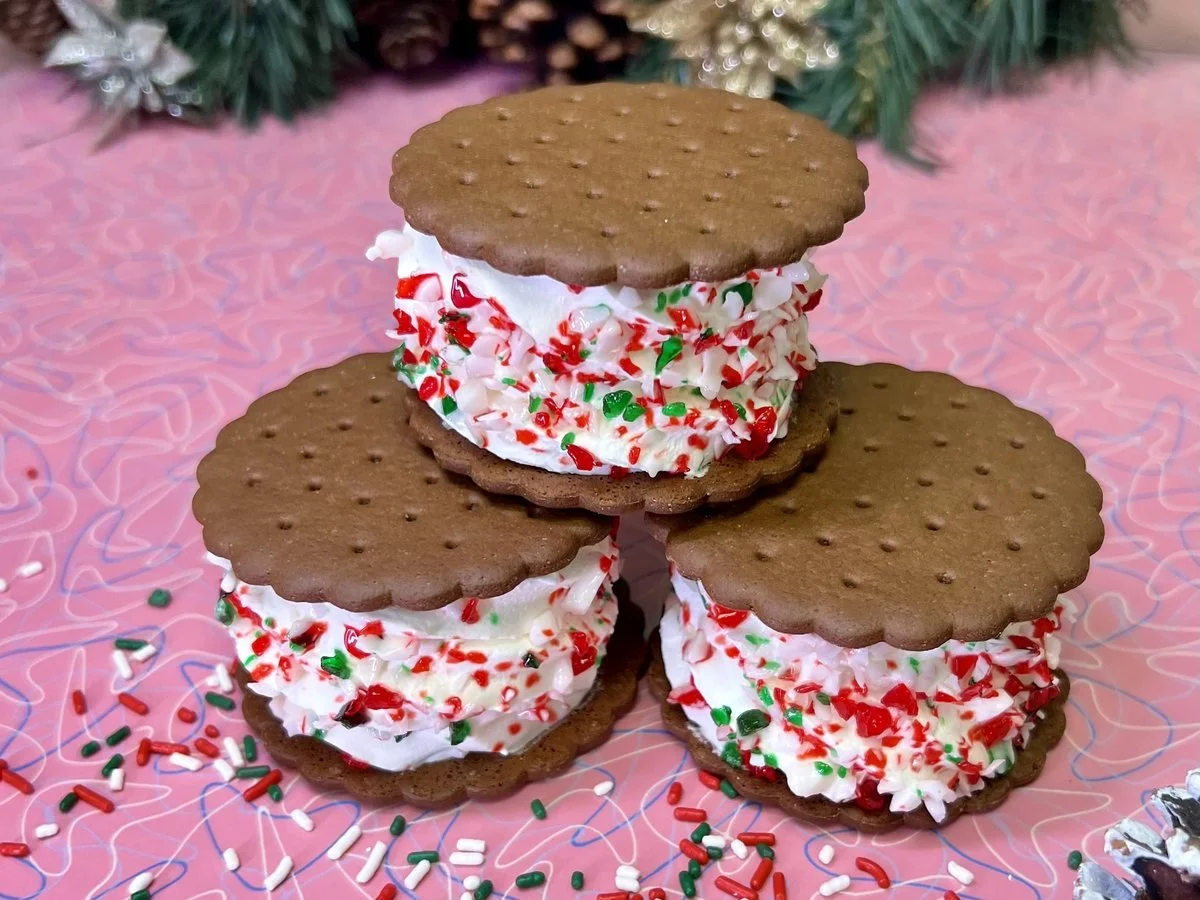 Three ice cream sandwiches with scalloped chocolate cookies, rolled in red, white and green candy and stacked on a pink table with pine boughs and pinecones behind, at Merla-Mae Ice Cream in London, Ontario