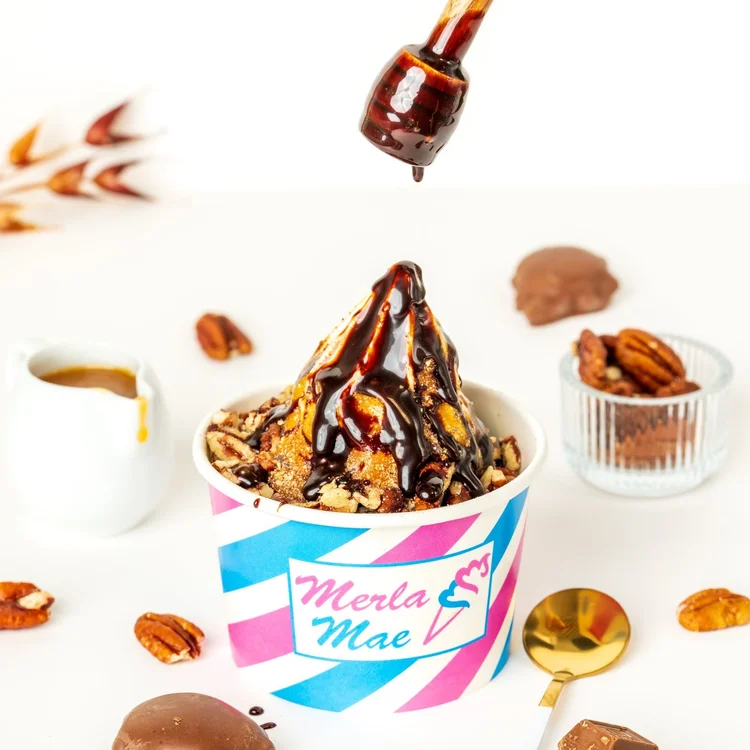 Chocolate sauce drizzling from a wooden dipper onto a Turtle sundae with caramel and chopped pecans in a striped Merla-Mae cup, with chocolate turtles and pecans around it, at Merla-Mae Ice Cream in London, Ontario