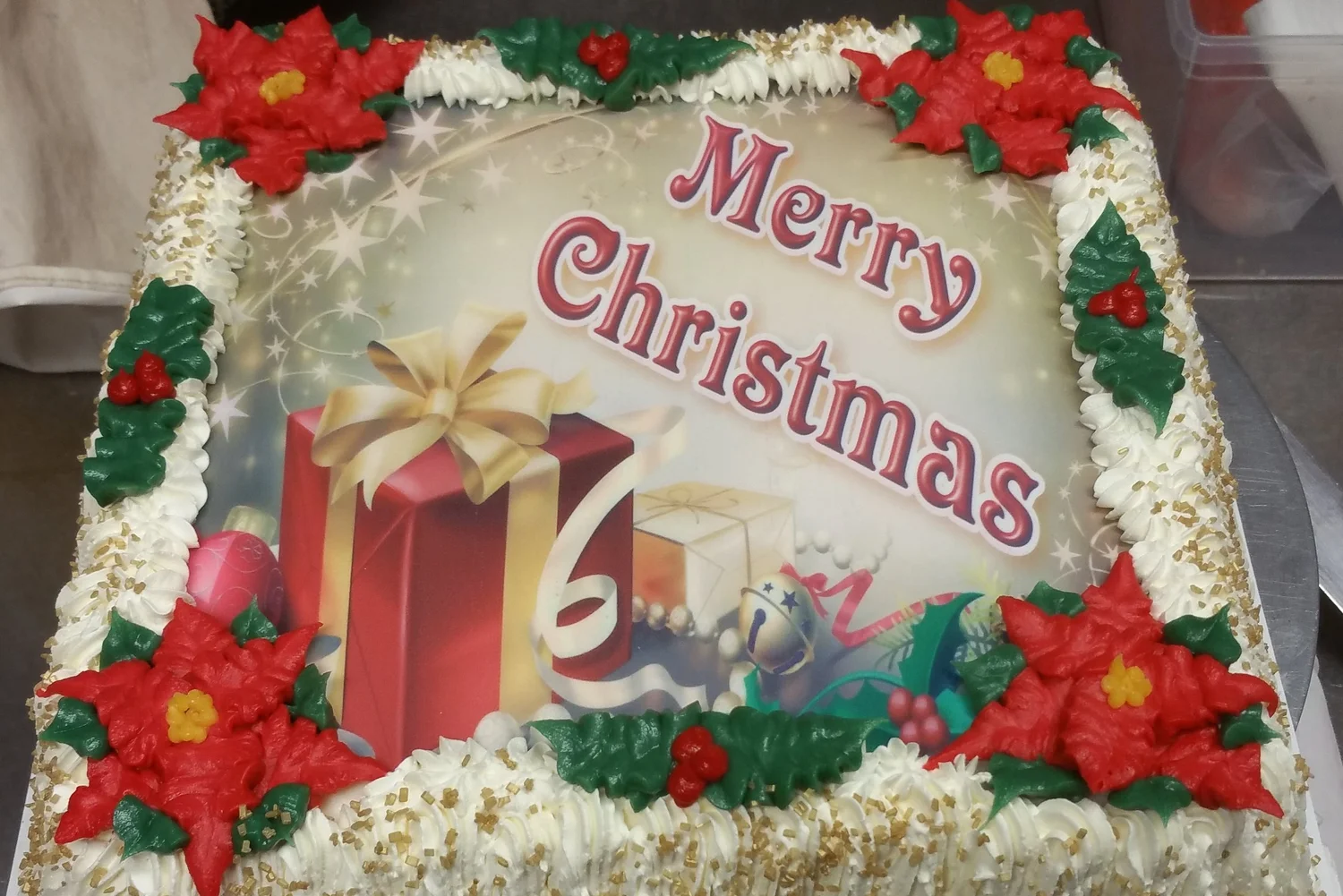 Merry Christmas ice cream cake with a printed gift-box picture, red icing poinsettias, green holly and gold sugar on white piped trim, at Merla-Mae Ice Cream in London, Ontario