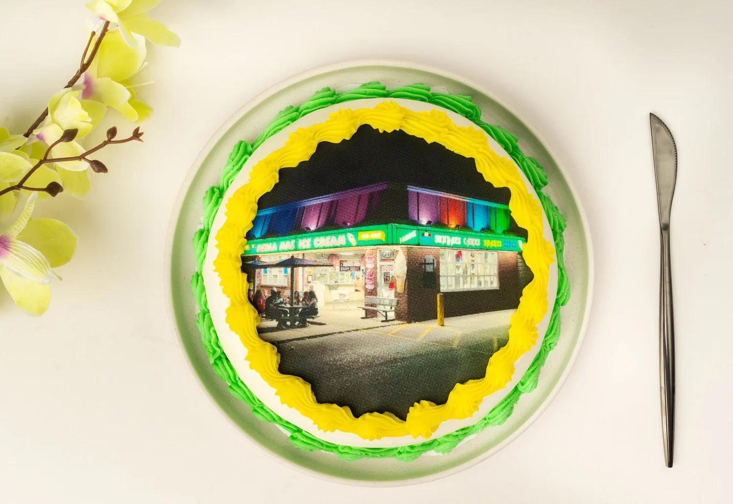 Round ice cream cake topped with an edible printed photo of the ice cream shop lit up at night, framed by yellow and green piped borders, beside a yellow orchid sprig and a cake knife, at Merla-Mae Ice Cream in London, Ontario