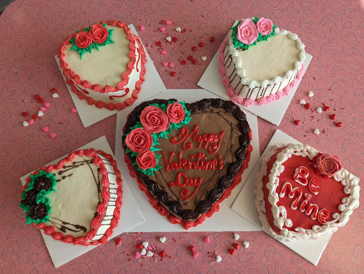 Five heart-shaped Valentine&rsquo;s ice cream cakes with icing roses, one reading Happy Valentine&rsquo;s Day and one reading Be Mine, on a pink boomerang-pattern table with heart sprinkles at Merla-Mae Ice Cream in London, Ontario