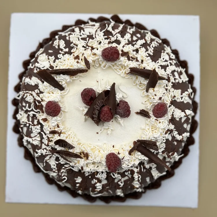 Round ice cream cake seen from above, with a white centre, whole raspberries, dark chocolate shards, white chocolate curls and a dark and white chocolate drizzle inside a chocolate shell border, at Merla-Mae Ice Cream in London, Ontario