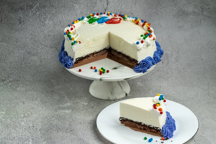 Ice cream cake on a marble cake stand with a wedge cut out to show a vanilla top over a chocolate base, edged with blue rosettes and rainbow candies, and a slice on a plate in front, at Merla-Mae Ice Cream in London, Ontario