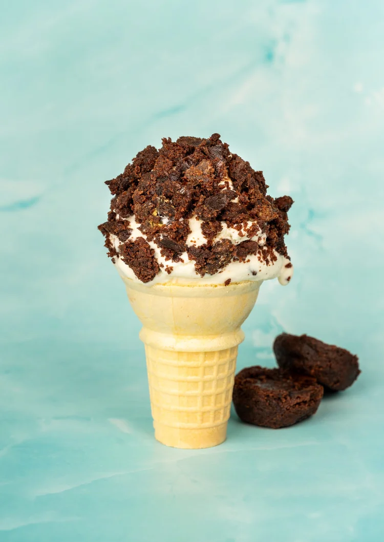 Scoop of ice cream rolled in brownie crumbs on a cake cone, with brownie pieces beside it, at Merla-Mae Ice Cream in London, Ontario