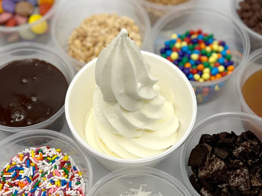 A tall, peaked swirl of vanilla soft-serve in a white cup, surrounded by open tubs of toppings: rainbow sprinkles, crushed chocolate cookies, chocolate sauce, a golden sauce, candy-coated chocolates and a crunchy crumble, at Merla-Mae Ice Cream in London, Ontario