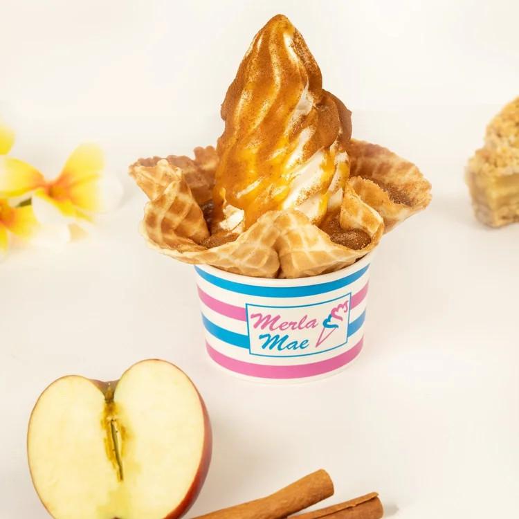 Soft-serve drizzled with caramel and dusted with cinnamon in a waffle bowl, in a striped Merla-Mae cup beside a halved apple and cinnamon sticks, at Merla-Mae Ice Cream in London, Ontario
