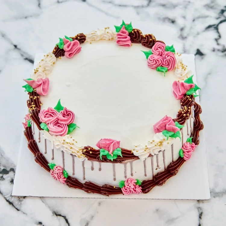 Round white ice cream cake with piped pink roses and green leaves, a chocolate border with gold sprinkles and chocolate drips down the sides, on white marble at Merla-Mae Ice Cream in London, Ontario