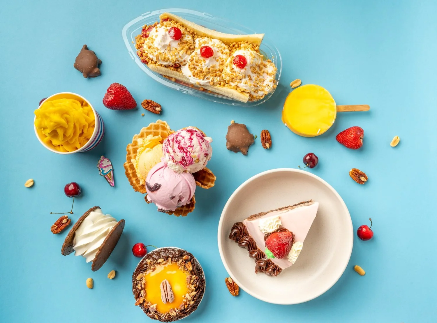 A banana split, scoops of ice cream in a waffle bowl, an ice cream sandwich, a swirled orange treat in a cup, a dipped bar on a stick and a slice of strawberry-topped cake spread out on a blue table at Merla-Mae Ice Cream in London, Ontario
