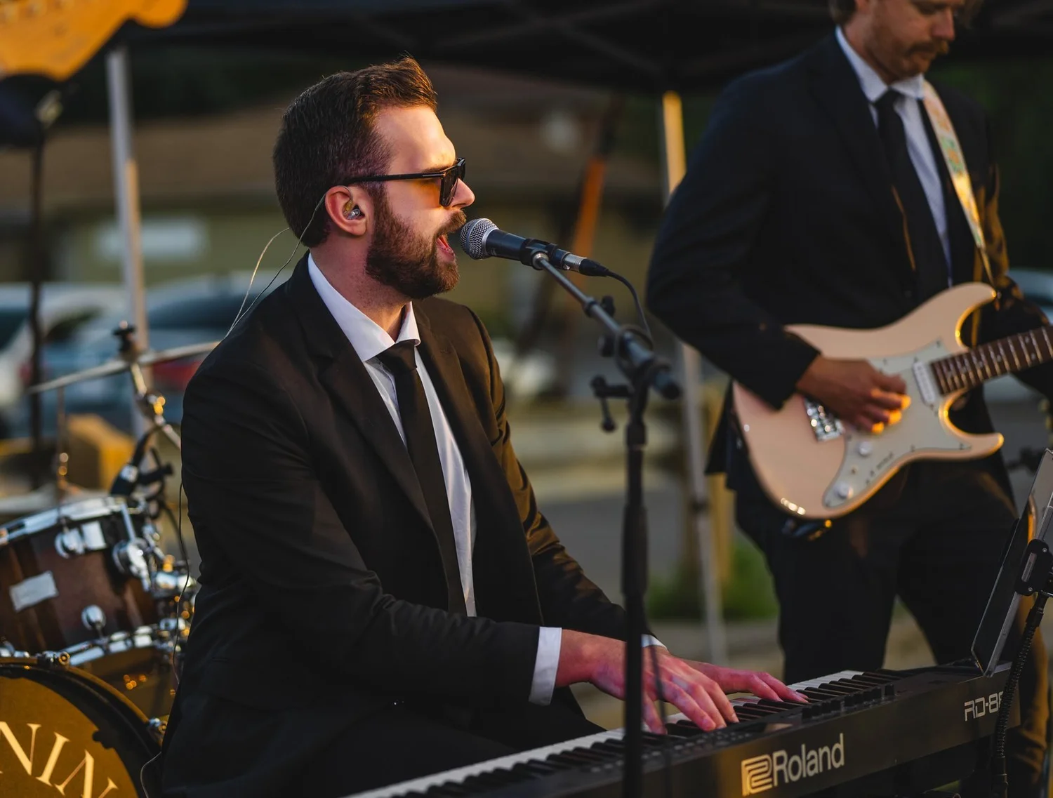 Turning Corners' singer at the keyboard with a guitarist behind him, both in suits, at National Ice Cream Day 2025, Merla-Mae Ice Cream in London, Ontario
