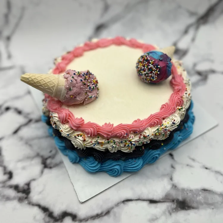 Round ice cream cake with a pink piped border and blue base, topped with two sprinkle-covered ice cream cones, at Merla-Mae Ice Cream in London, Ontario