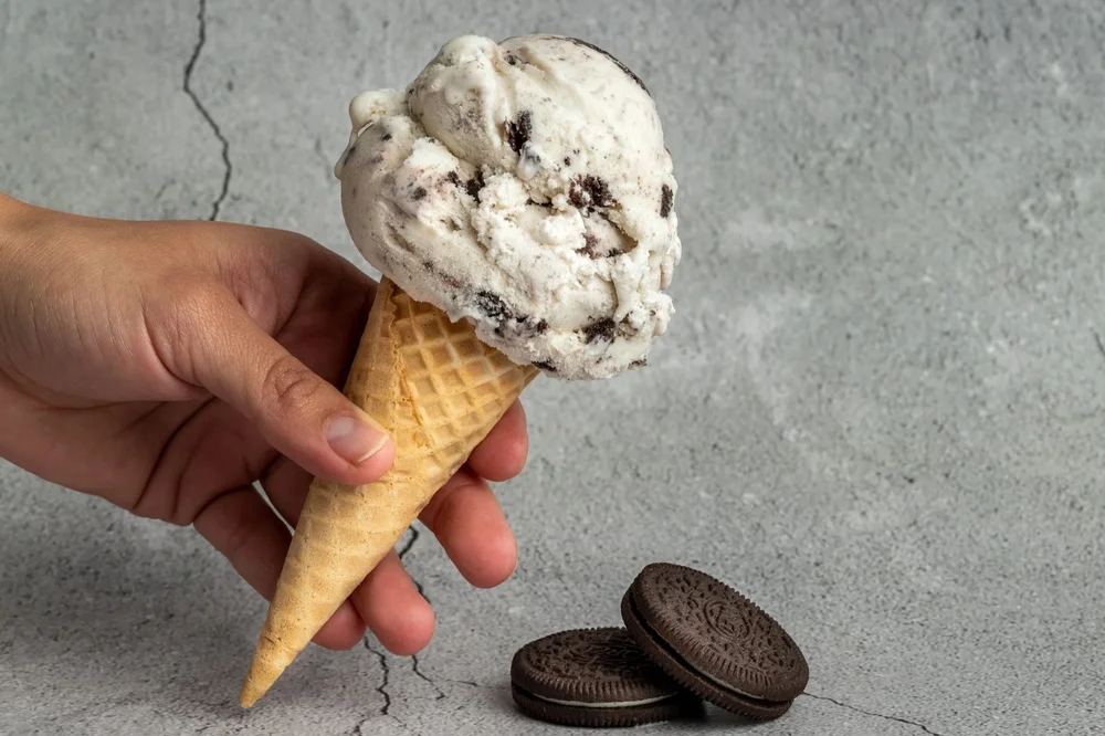 A hand holding a cone with a scoop of white ice cream full of dark cookie pieces, next to two chocolate sandwich cookies, at Merla-Mae Ice Cream in London, Ontario