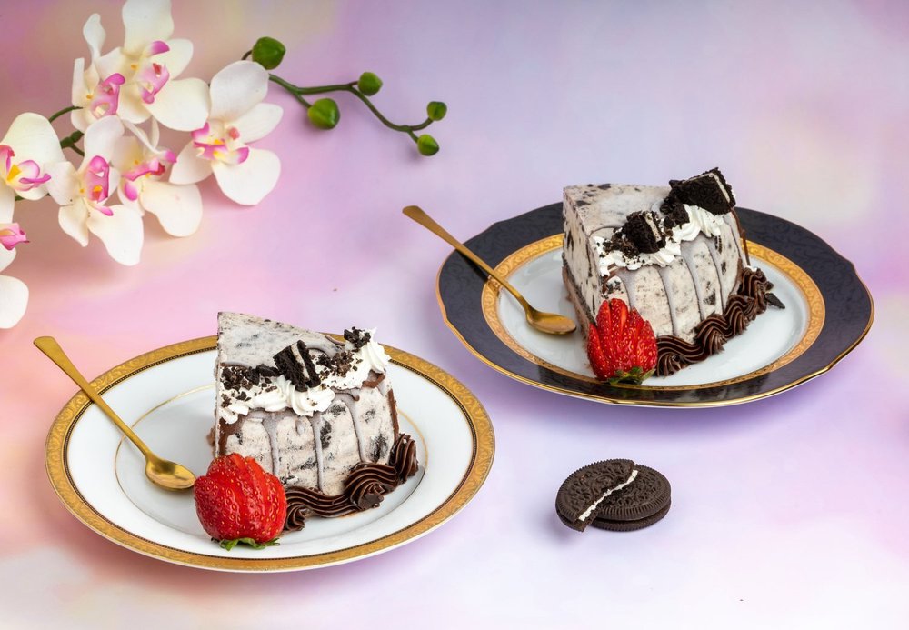 Two slices of Oreo ice cream cake topped with whipped cream and Oreo cookie pieces, each with a fresh strawberry, on gold-rimmed china plates with gold spoons beside white and pink orchids, at Merla-Mae Ice Cream in London, Ontario