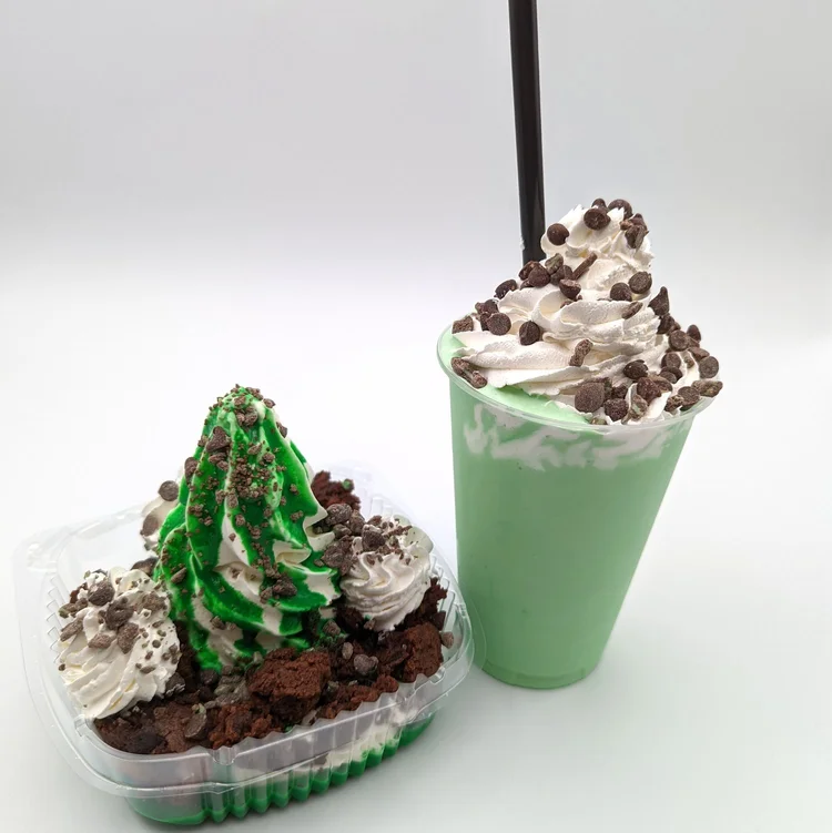 Green and white swirled soft-serve sundae on brownie pieces with whipped cream and chocolate chips, beside a mint-green shake topped with whipped cream and chocolate chips, at Merla-Mae Ice Cream in London, Ontario