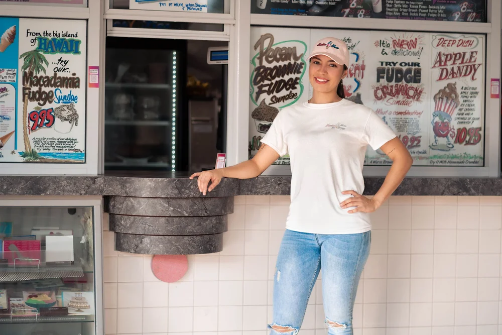 Smiling Merla-Mae team member in front of the hand-painted sundae signs inside Merla-Mae Ice Cream in London, Ontario