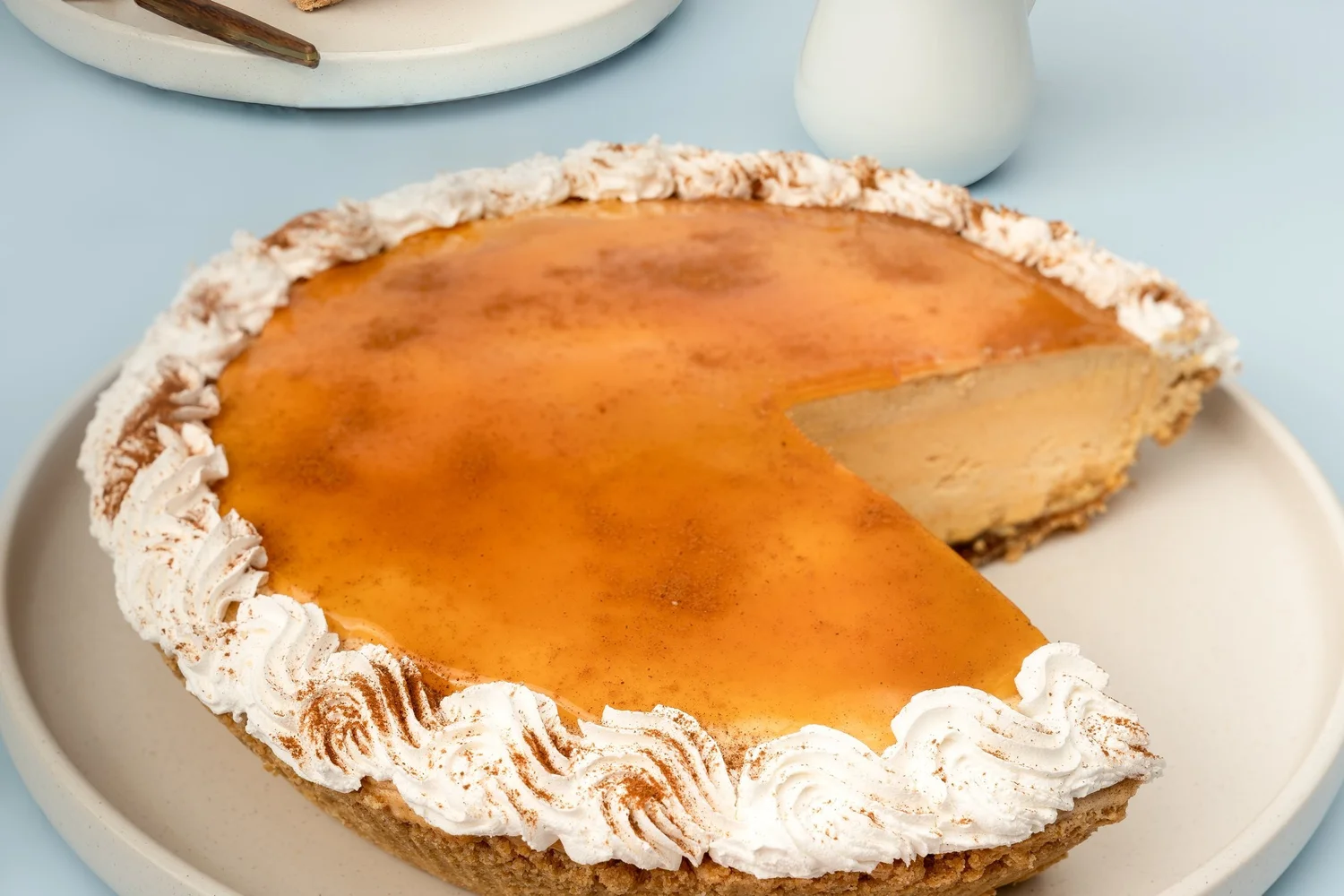 Pumpkin ice cream pie with a glossy amber top and a rim of whipped cream rosettes dusted with cinnamon, one wedge cut to show the filling, on a cream plate against pale blue, at Merla-Mae Ice Cream in London, Ontario